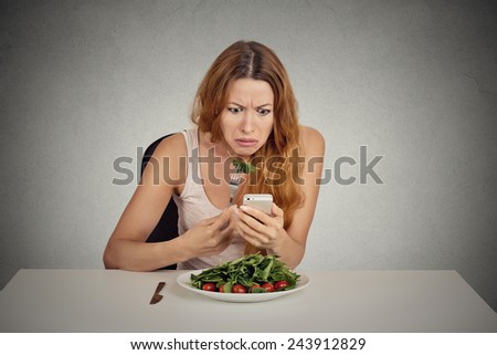 portrait young girl eating green salad looking at phone seeing bad news or photos annoyed  confused disappointed face expression isolated on grey wall background
