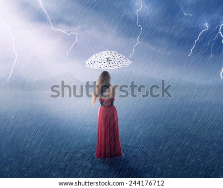 Beautiful young woman in red dress under umbrella on countryside flooded field on rainy day with thunderstorm cloudy sky landscape background. Safety, financial security, risk, life insurance concept