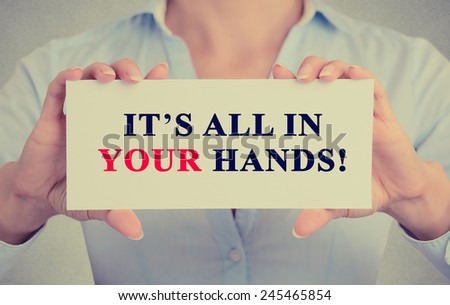 Image of businesswoman hands holding white card with It's All in Your hands! Motivating message sign isolated on grey wall office background.