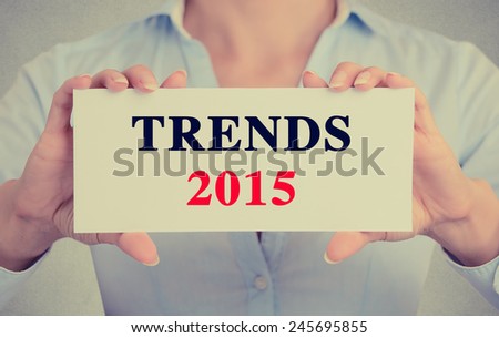 Businesswoman hands holding white card sign with trends 2015 text message isolated on grey wall office background. Retro instagram style image