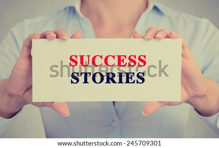 Businesswoman hands holding white card sign with success stories text message isolated on grey wall office background. Retro instagram style image. Inspiration progress concept