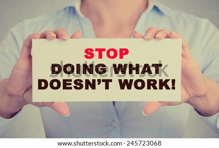 Businesswoman hands holding white card sign with Stop Doing What Doesn\'t Work text message isolated on grey wall office background. Retro instagram style image