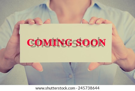 Businesswoman hands holding white card sign with coming soon text message isolated on grey wall office background. Retro instagram style image