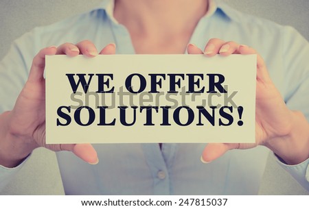 Businesswoman hands holding white card sign with we offer solutions! text message isolated on grey wall office background. Retro instagram style image