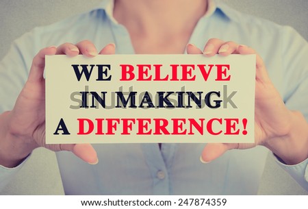 Businesswoman hands holding white card sign with We Believe in Making a Difference text message isolated on grey wall office background. Retro instagram style image