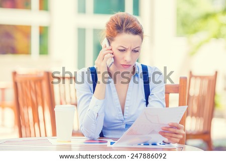 Closeup portrait upset sad, skeptical, unhappy, serious woman talking on phone holding looking at documents outside corporate office background. Negative human emotion face expression feeling reaction