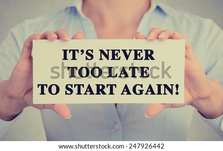 Businesswoman hands holding white card sign with It's never too late to start again text message isolated on grey wall office background. Retro instagram style image