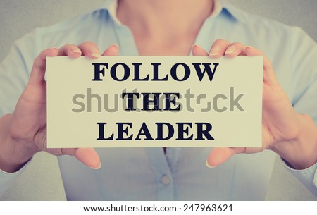 Businesswoman hands holding white card sign with follow the leader text message isolated on grey wall office background. Retro instagram style image