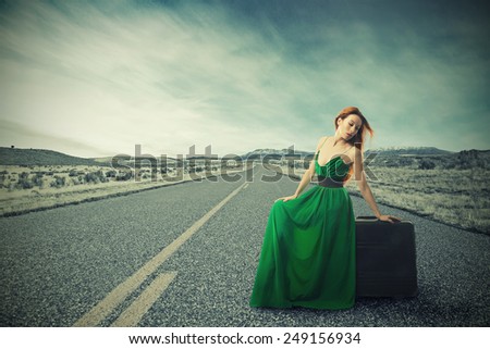 Woman sitting on a suitcase on a countryside road waiting for a ride