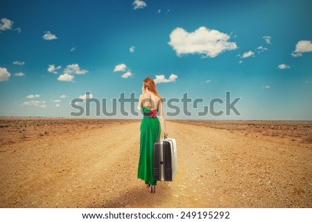 Beautiful woman walking through a desert road talking on mobile phone carrying big suitcase isolated on blue cloudy sky background. Embrace challenge unknown new life loneliness concept. Long journey