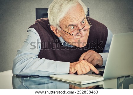 Elderly old man using computer sitting at table isolated on grey wall background. Senior people and technology concept