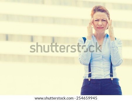 Portrait stressed worried young woman corporate employee having headache migraine  isolated outdoors on corporate office windows building background. Negative human emotions face expression