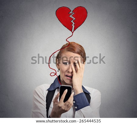 Sad woman with broken heart looking at her mobile phone isolated on grey wall background