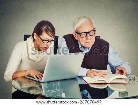 Portrait young woman working on laptop and older senior mature grandpa man reading from book on table isolated gray wall background. Generation differences and gap technology concept