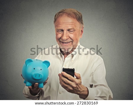 Portrait happy senior man holding piggy bank looking at smart phone isolated grey background. Financial savings banking concept, customer satisfaction contract agreement. Positive face expression