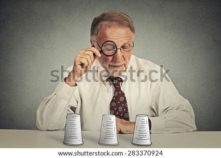 Portrait senior elderly  business man with glasses sitting at table desk looking through magnifying glass wondering playing conjuring trick game isolated grey wall background. Gambling risk concept