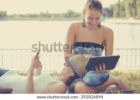 Two funny happy young women friends laughing browsing sharing watching social media videos blogs on smart phone pad computer laying outdoors on green meadow on sunny summer day