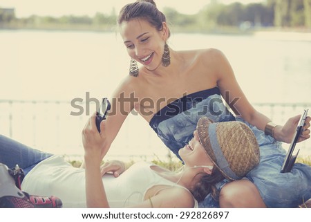Two funny happy young women friends laughing browsing sharing watching social media videos blogs on smart phone pad computer laying outdoors on green meadow on sunny summer day