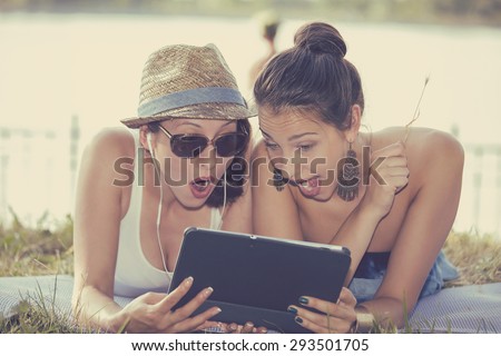 Closeup portrait two surprised girls looking at pad discussing latest gossip news. Young shocked funny women friends reading sharing social media news on mobile pad computer outdoors in park