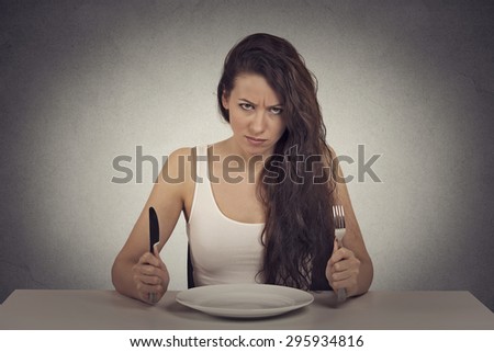 Young skeptical dieting woman tired of diet restrictions looking at camera sitting at table with empty plate with fork and knife. Human face expression emotion. Nutrition concept