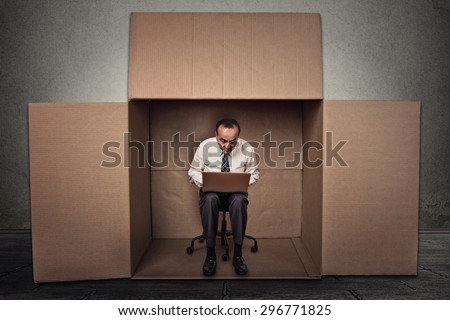 Limitations poor communication of corporate life. Portrait corporate middle aged man working on laptop sitting on chair inside carton box in empty office room