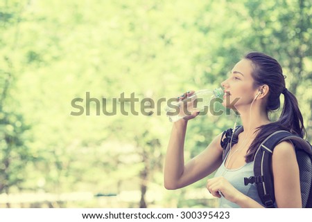 Side profile attractive happy young woman hiker with backpack drinking bottled water walking on a country forest trail landscape on a summer hot day