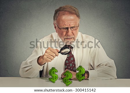 penny pincher. Senior skeptical business man looking through magnifying glass at dollar sign symbol isolated gray background. Economy financial wealth success concept. Ponzi scheme investigation