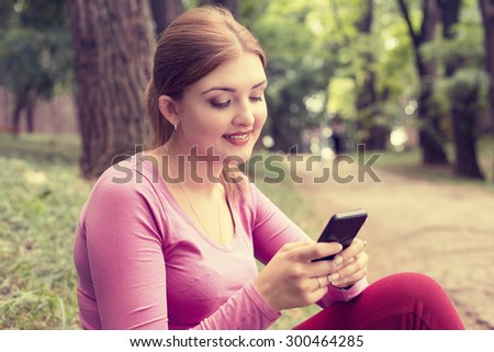 Closeup portrait, happy, cheerful, young woman excited by what she sees on cell phone sitting outdoors in park. Facial expression, reaction. Smiling girl sending text message from her mobile