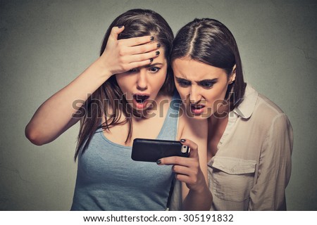 Two shocked women displeased young girls looking at mobile phone seeing bad news message or photos with disgusting emotion on face isolated on gray wall background. Human emotion, reaction, expression