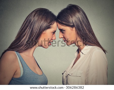 Side profile unhappy young female friends looking at each other standing head to head on gray wall background. Friendship difficulties, problems at work concept