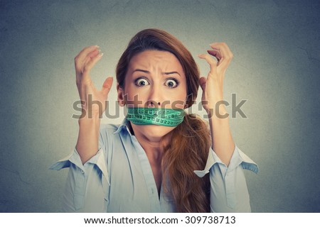 Diet restriction and stress concept. Portrait of young frustrated woman with a green measuring tape around her mouth isolated on gray wall background. Face expression emotion