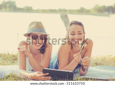 Two funny happy young women friends laughing browsing sharing watching social media videos blogs on pad computer laying outdoors on green meadow on sunny summer day