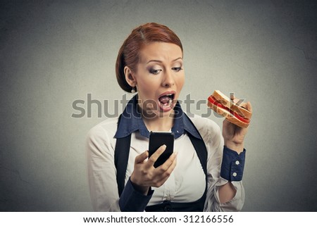 young corporate business woman reading news message on smart phone holding eating sandwich isolated on gray background. Human face expression.