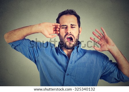 Young sleepy man yawning stretching arms back isolated on gray wall background. Sleep deprivation, burnout, laziness concept