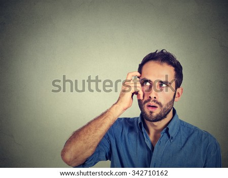 Closeup portrait young man scratching head, thinking deeply about something, looking up, isolated on grey wall background. Human facial expression, emotion, feeling, sign body language