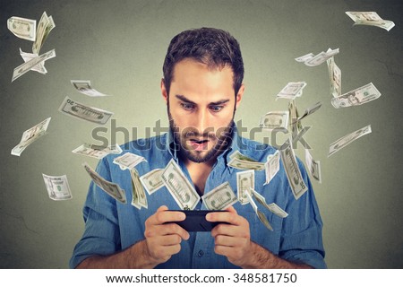 Technology online banking money transfer, e-commerce concept. Shocked young man using smartphone with dollar bills flying away from screen isolated on gray wall office background.