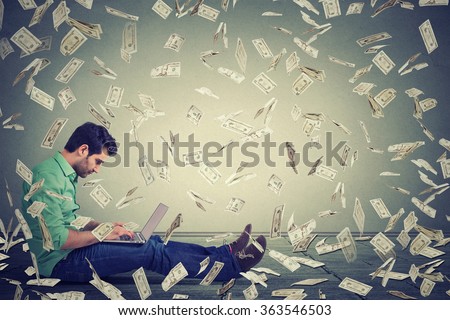 Young man using a laptop sitting on a floor building online business making money dollar bills cash falling down. Money rain. Beginner IT entrepreneur success economy concept
