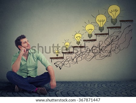 Happy man sitting on a floor in his office dreaming of business education success, promotion, company growth isolated gray wall texture background. Handsome guy looking up at growing up light bulbs