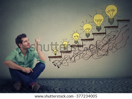 Happy man sitting on floor in his office pointing at business education success, promotion, company growth isolated gray wall texture background. Handsome guy looking up at growing up light bulbs