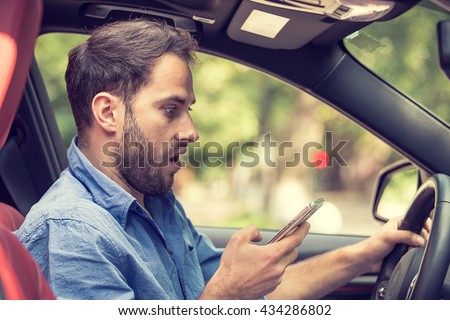 Man sitting in car with mobile phone in hand texting while driving. Distracted shocked guy checking his smart phone not paying attention at road annoyed by bad text message email outdoors background