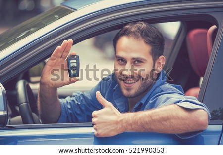 Happy, smiling, young man sitting in his new blue car showing keys thumbs up isolated outside dealership lot. Personal transportation purchase concept