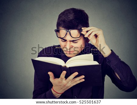 Closeup young man with eye glasses trying to read book, having difficulties seeing text, has sight problems. Eyesight issues concept. Human face expression