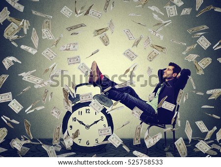 Happy young business man relaxing sitting in his office under money rain making dollar bills cash falling down. Stress free time management good earnings profit concept