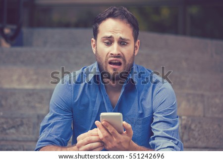 Anxious upset young scared man looking at phone seeing bad news or text message sitting on stairs outside corporate building. Human emotion, reaction, expression