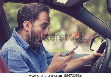Man sitting inside car with mobile phone in hand texting while driving. Shocked guy checking his smartphone not paying attention at road stunned by bad text message email outdoors background