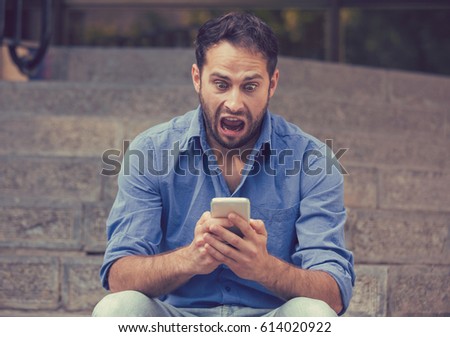Shocked upset young man looking at his mobile phone seeing bad news or reading text message sitting on stairs outside corporate building. Human emotion, reaction, expression