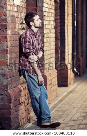 Pensive young man leaning against the brick wall and waiting for someone, he is wearing checked shirt and jeans