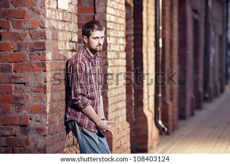 Pensive young man leaning against the brick wall and waiting for someone, he is wearing checked shirt and jeans