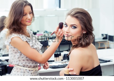 Make-up artist applying powder with a brush on model\'s cheeks, selective focus on model looking at camera