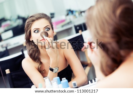 Young beautiful woman making faces and joking  with a make-up brush in front of a mirror